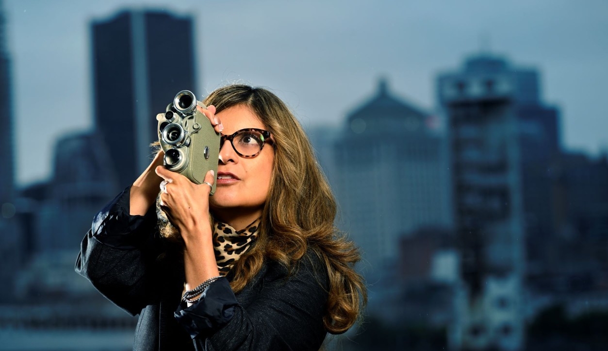 Woman with long, brown hair wearing a black jacket and tortoiseshell glasses holds a video camera to her eye with a city skyline in the backgound. 