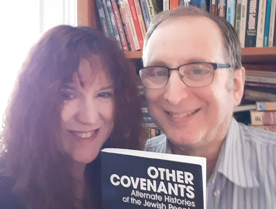 woman with long, curly hair and man with short, brown hair wearing black-rimmed glasses hold up a book. 