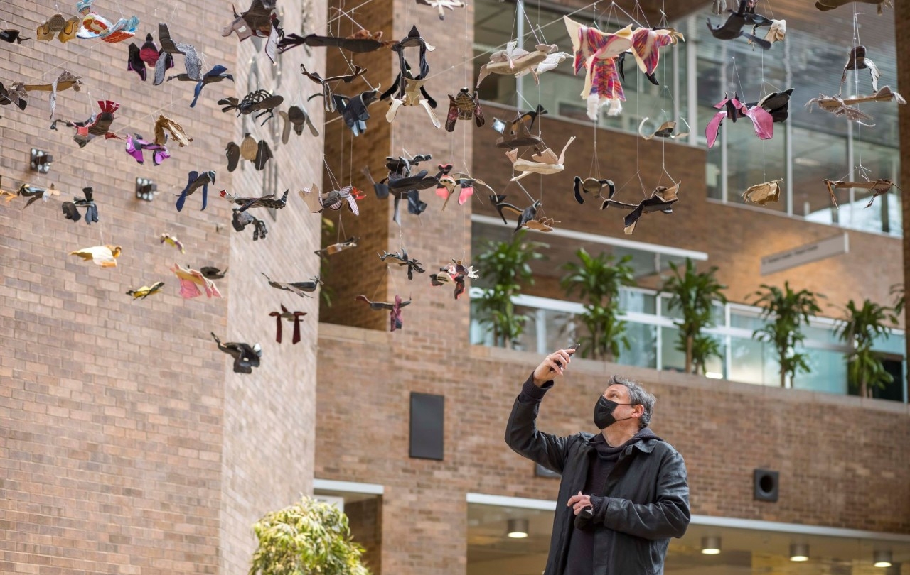 A man wearing a mask holds a camera as he takes photo of hanging paper birds with an apartment building in background