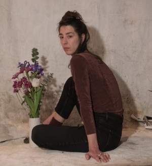 Portrait of a woman with long, dark hair, wearing brown velvet top and black pants, pictured with a vase of flowers