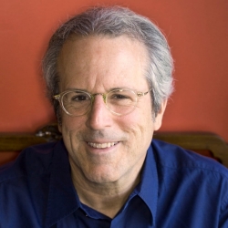 René Balcer wears glasses and a blue shirt. He is sitting in front of a red background.