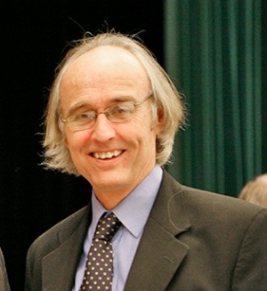 Portrait of a man with light grey hair, wearing glasses, suit blazer and blue dress shirt with polka-dot tie