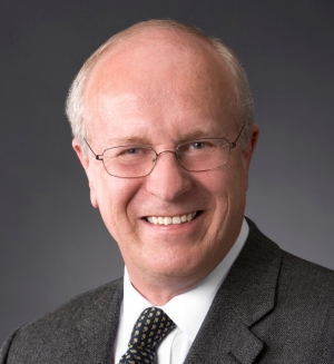 Portrait of a smiling man against a dark backdrop, wearing glasses and a dark blazer over a white shirt and dark tie