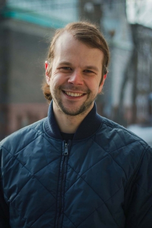 A man with light brown hair and beard smiles, wearing a navy blue zip-up jacket.