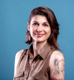 A smiling woman with short hair in front of a blue background is wearing a brown sleeveless shirt and has tattoos on her left arm
