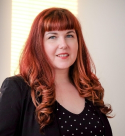 A woman with long red hair and bangs smiles at the camera. She is wearing a black shirt with white polka dots under a black blazer