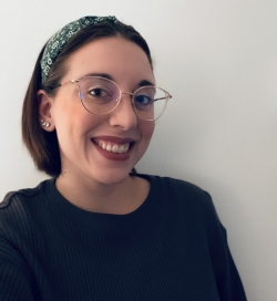 A smiling woman with short brown hair pulled back with a green headband is wearing glasses and a dark shirt