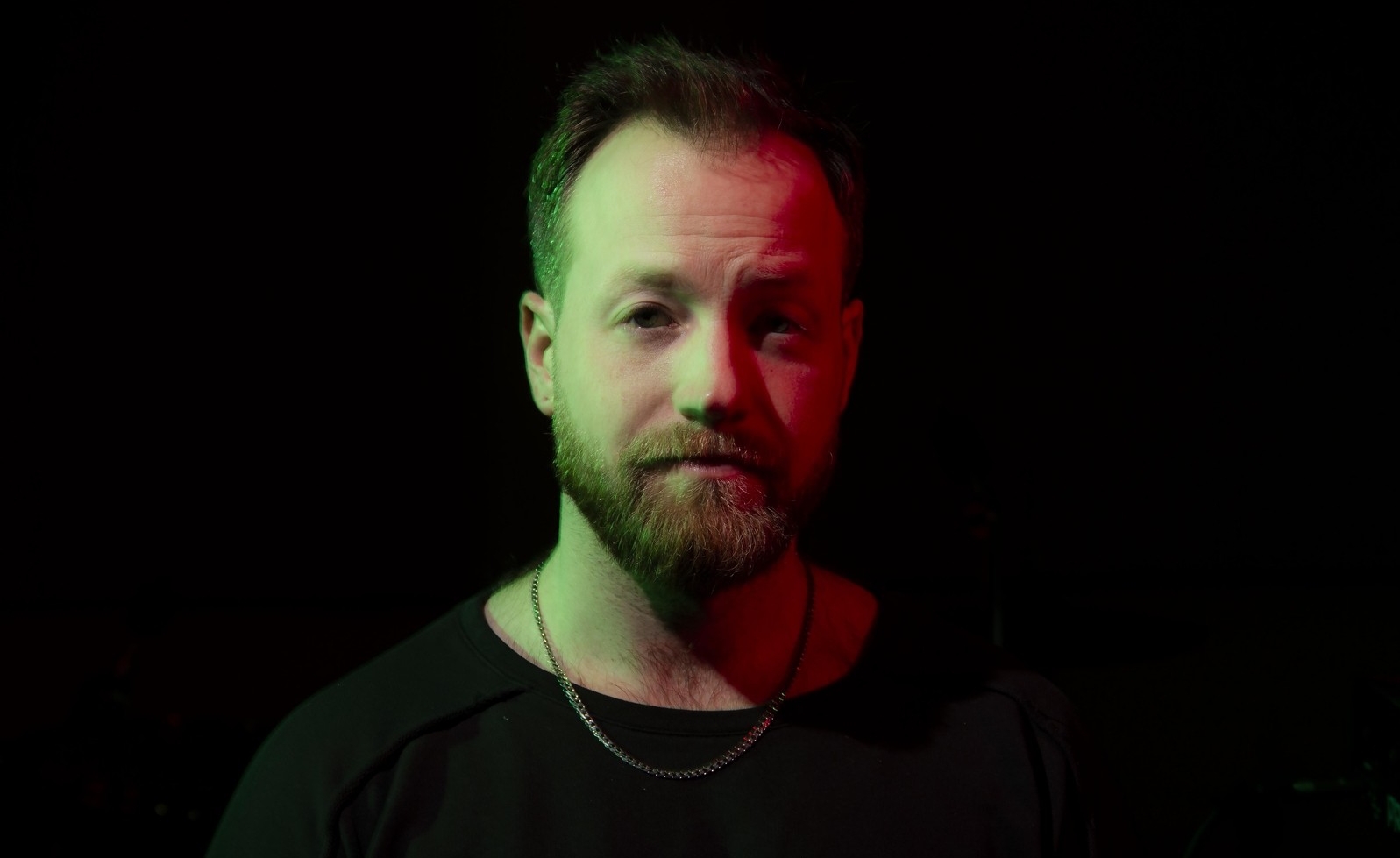 Headshot of a man with short light brown hair and a beard, wearing a black shirt, stands in front of a black backdrop