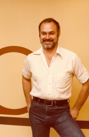 A photo of a man in 1979 with short, dark hair and beard smiling as he poses wearing a white collard shirt and blue jeans