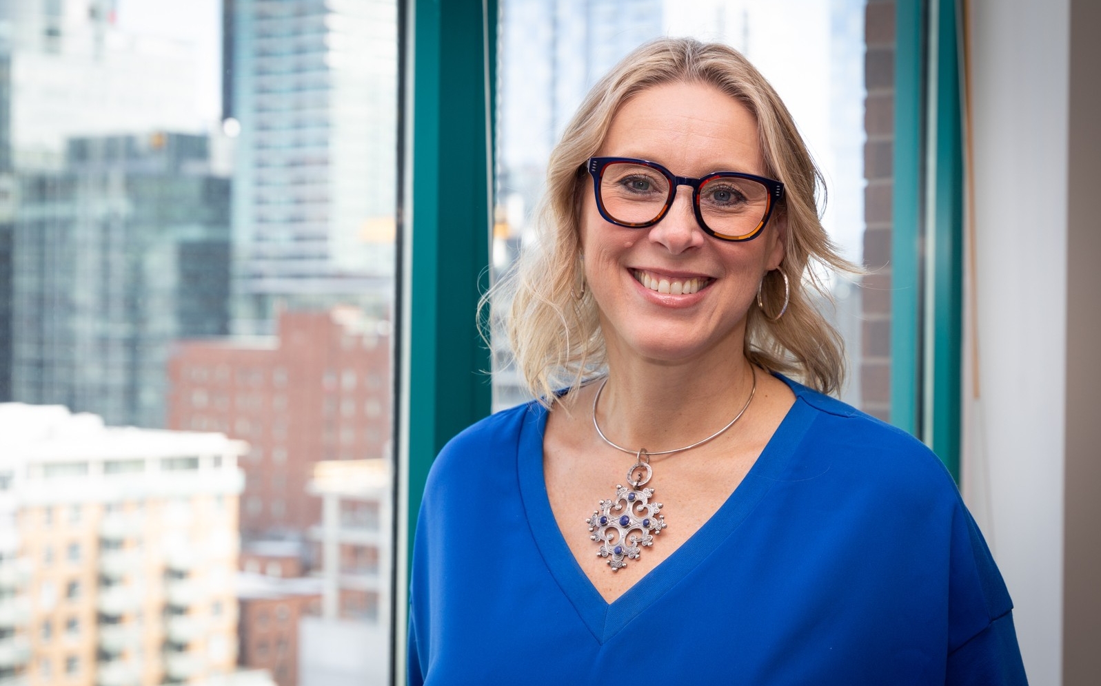 A photo of a woman wearing a royal blue v-neck sweater and necklace smiles in front of a window with buildings in the background. She has shoulder length blonde hear and is wearing dark framed glasses