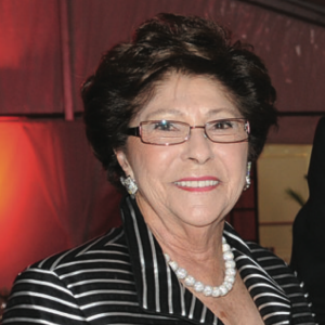 A woman with short brown hair smiles. She is wearing glasses, a pearl necklace and a black blazer with white stripes