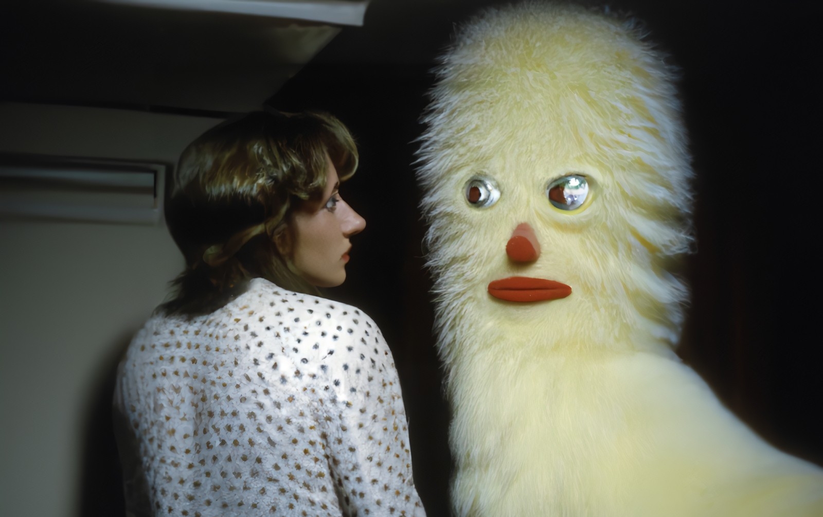 An AI-generated image of a woman with short brown hair looking at a life-size fluffy yellow figure that has big eyes, a red nose and red mouth