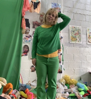 Image of a woman with short blonde hair wears a grean shirt and green pants. She is in a studio surrounded by stuffed toys in front of a white wall and green curtain