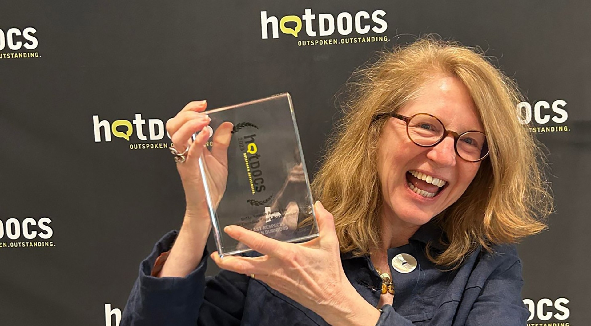 A blonde women with glasses smiles and holds an award aloft