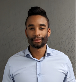 A man with black hair and light blue dress shirt faces the camera; he is standing in front of a grey wall
