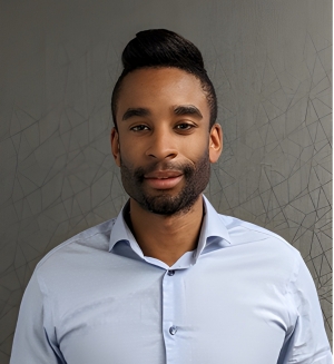 A man with black hair and light blue dress shirt faces the camera; he is standing in front of a grey wall