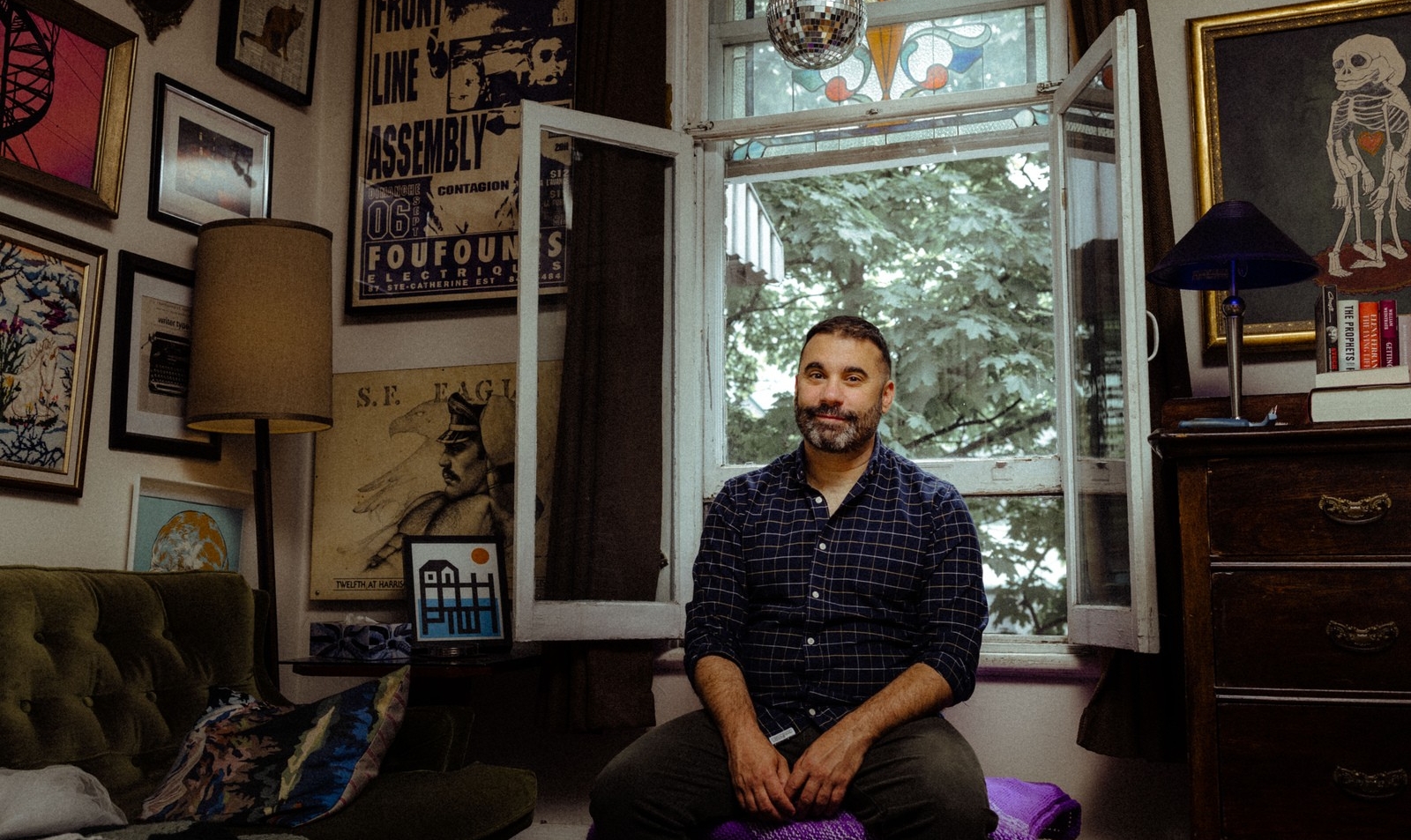 Image of a man with short dark hair and beard sitting in front of an open window in a room with walls covered in photographs and art
