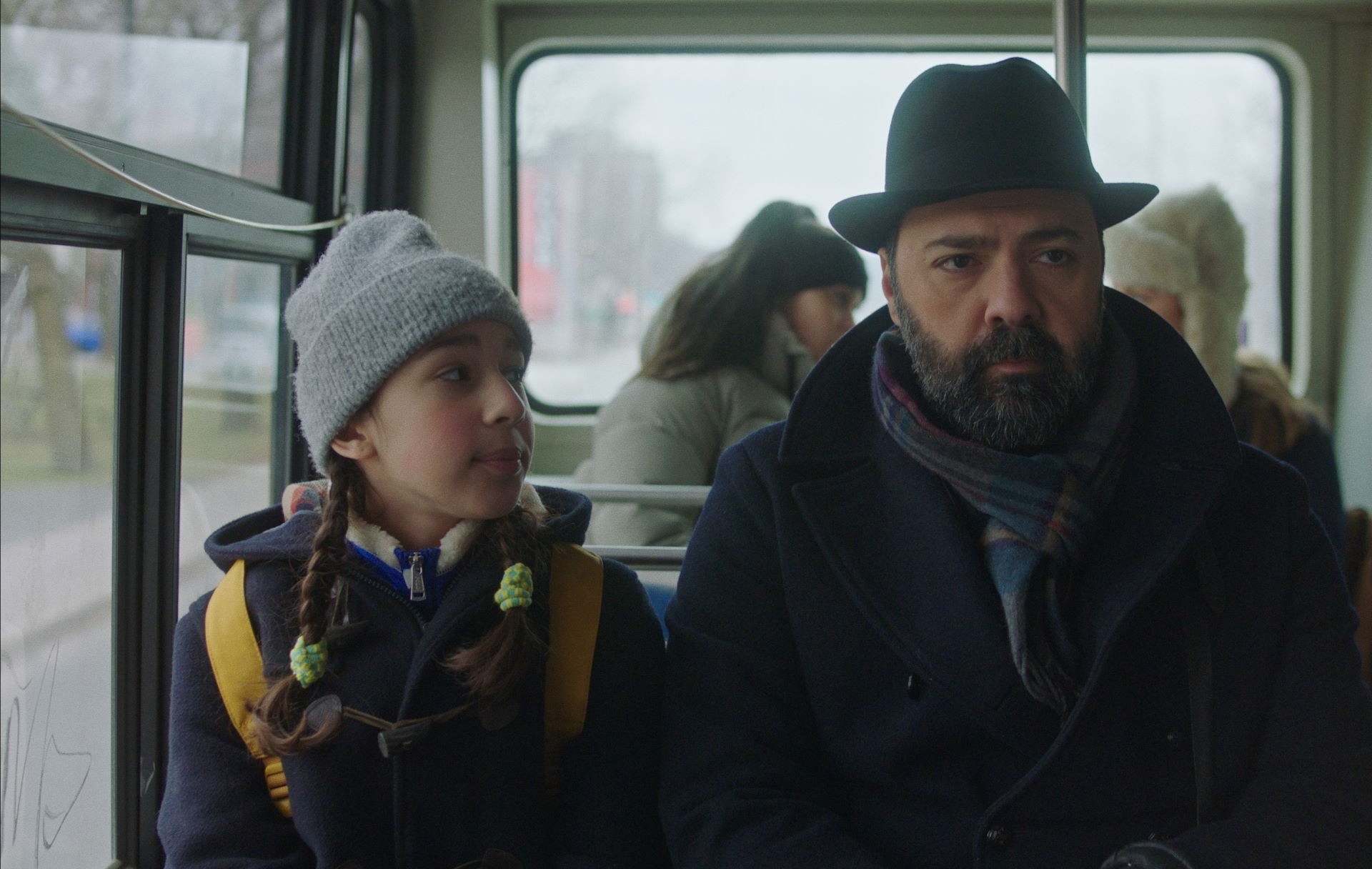 A girl and man dressed in winter clothes sit on a bus. The girl wears a grey toque and looks up at the man who wears a black hat.