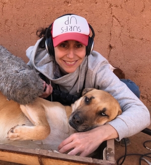 A woman wears a red and white hat, headphones, and a grey hoodie. She has one arm wrapped around a large, brown dog, and the other holds a microphone.