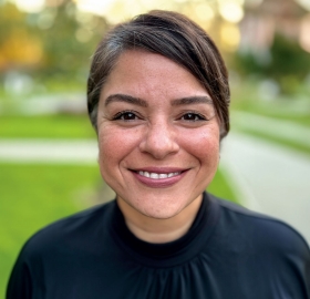 Close-up portrait of Puja Chaubey, a woman with a warm smile, medium-length dark hair parted to the side, and clear skin. She is wearing a black high-neck top and subtle makeup with natural-toned lipstick. The background is softly blurred with hints of greenery, suggesting an outdoor setting.