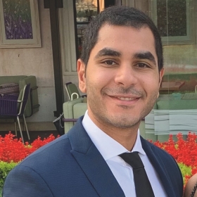 Portrait of Karim Refaat, a man with a friendly smile, sporting short dark hair and light stubble. He is dressed in a sharp navy blue suit with a white shirt and black tie. In the background, there is an outdoor setting with a glass door, artwork on the wall, and a blur of green chairs and vibrant red flowers, suggesting a relaxed, yet elegant ambiance.