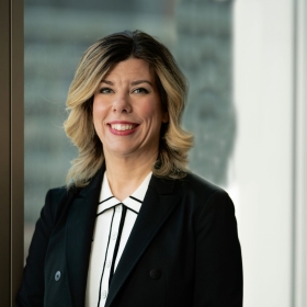 The image shows a professional portrait of Marie-Chantal Côté. She is smiling and appears confident. The setting seems to be an office or a corporate environment, indicated by the blurred background which suggests an interior with windows. She has shoulder-length wavy hair, styled neatly, and is wearing what appears to be a business suit with a black jacket and a white shirt with a black collar. 