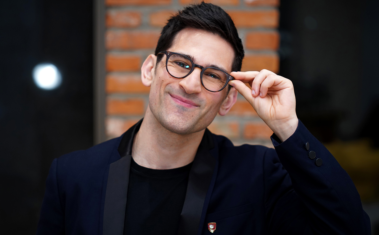 A man with dark hair is wearing a black blazer over black t-shirt. He is wearing black framed glasses and is touching them with his left hand.