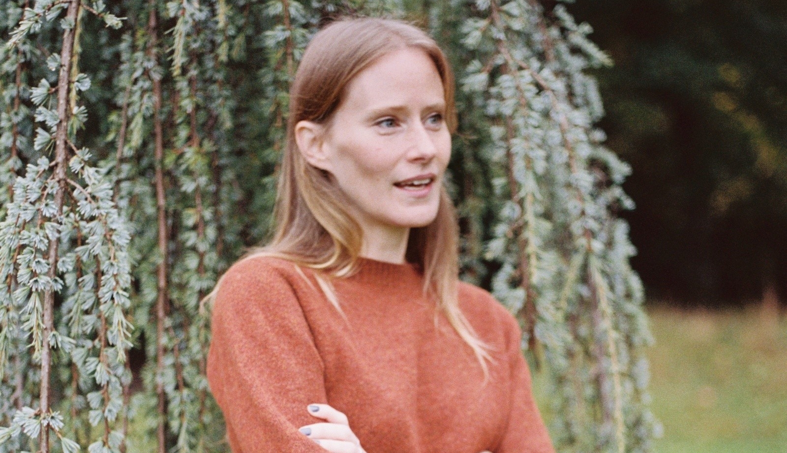 A woman with long blonde hair is wearing light red sweater, standing in front of a tree
