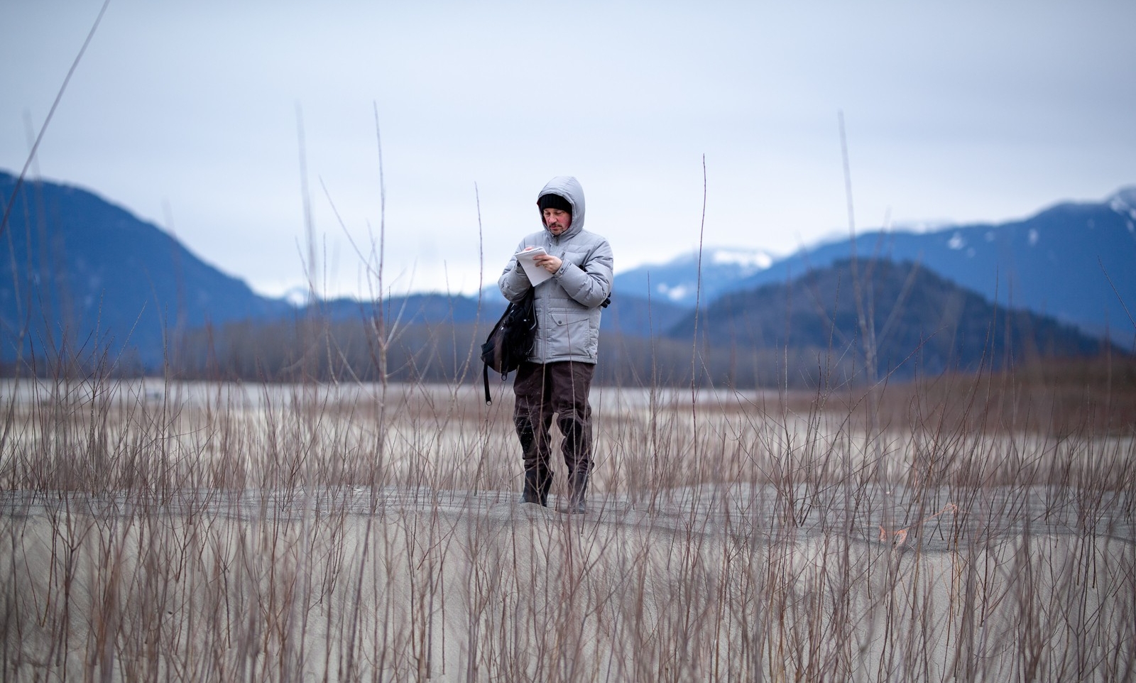 A man with a light grey hooded jacked and black pants stands in a field with snow-capped mountains behind him. He is taking notes on a pad of paper.