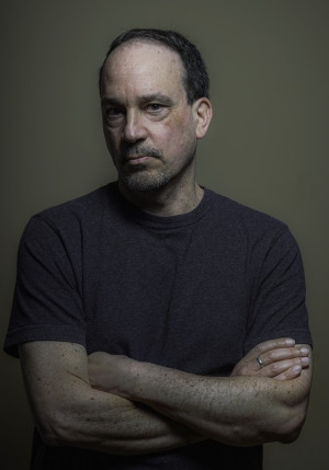A man with short dark hair and scruffy face stands with his arms folded. He is wearing a dark t-shirt and is standing in front of a dark background