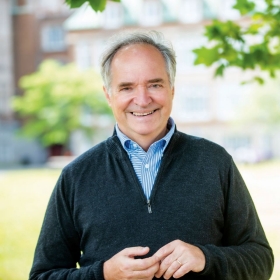 A middle aged gentleman stands for portrait taken outside with trees, grass and buildings faded in the background