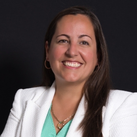 A woman with long brown hair in a white blazer and green shirts sits smiling for a portrait on a black background