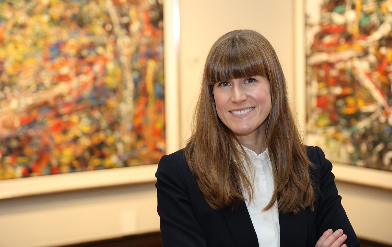A woman with long brown hair and bangs is wearing a dark blazer over a white collared shirt. She is smiling and is standing in front or paintings by Quebec artist Jean-Paul Riopelle.