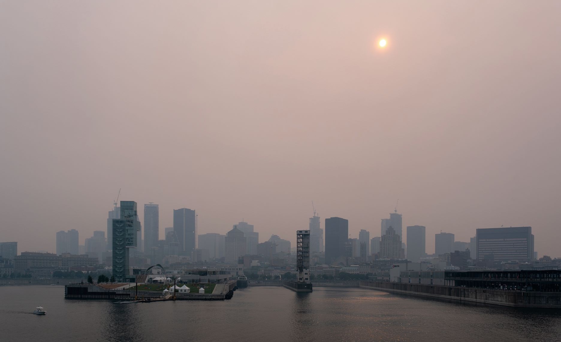 A hazy skyline of Old Montreal with muted colors, featuring the sun as a pale disc in a smoggy sky. The Saint Lawrence River is in the foreground reflecting the city's silhouette, and various buildings are visible, some under construction. There's a sense of calm despite the overcast atmosphere, with a small boat visible on the water.