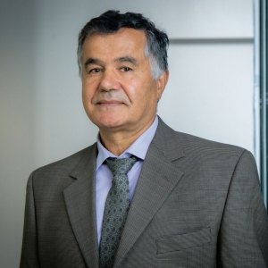 A portrait of a professional man with dark hair and a slight smile, wearing a gray suit, patterned tie, and a lavender shirt. He is standing in an office environment, with a blurred background featuring vertical window blinds to the right.