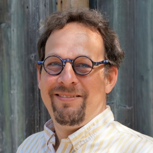A close-up portrait of a man with a friendly smile, wearing round-framed glasses. He has curly, dark hair with touches of gray and a short, neat beard. The man is dressed in a light-colored, striped button-down shirt, and the background features a rustic, weathered wooden fence that adds a textured backdrop to the image.