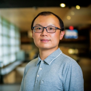 A portrait of a man wearing a light grey, collared shirt and glasses. He is looking directly at the camera with a subtle smile. The background is an indoor setting, softly focused with rows of frosted glass windows and warm lighting, which suggests a corporate or academic environment.