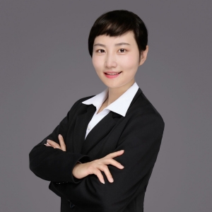 A professional portrait of a young woman with short hair, smiling at the camera. She is wearing a dark business suit with a white collared shirt and has her arms crossed in front of her, conveying confidence. The background is a plain, neutral grey, putting the focus on her.