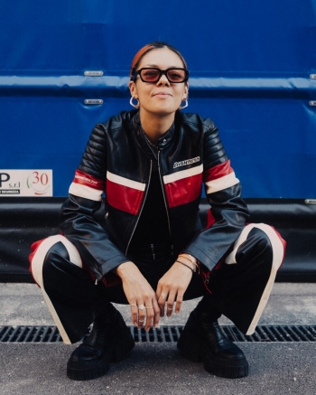 Portrait of a woman with dark hair, wearing silver earring and black-rimmed glasses, leather jacket and pants