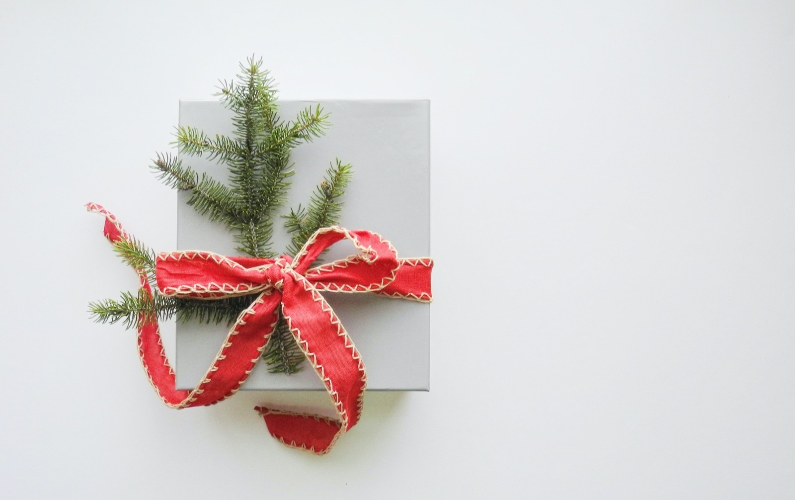 A small, light grey square box is tied with a red ribbon and small piece of pine. It sits against a white background