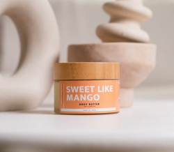 A light container with a wooden lid has as orange label with white text that says Sweet Like Mango body butter