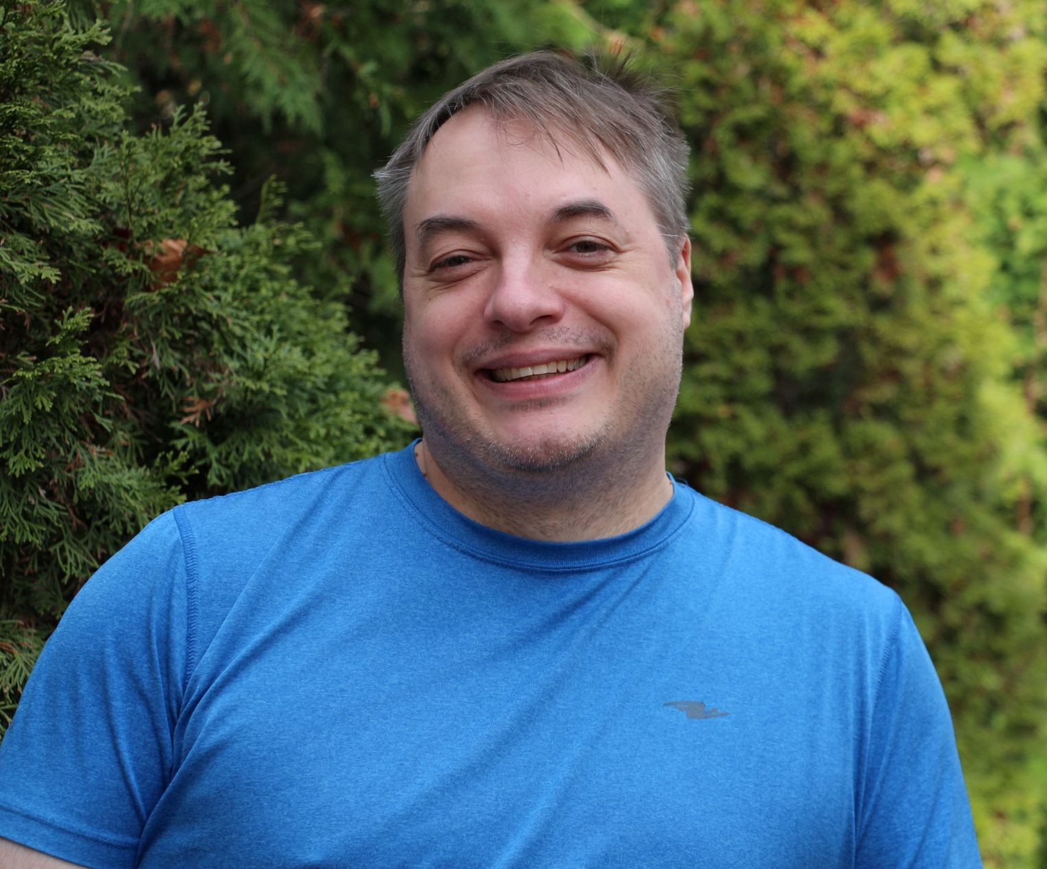 A man in a blue shirt smiling outside in front of green shrubs.
