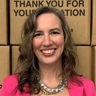 A woman with long brown hair wearing a collection of silver necklaces and a pink jacket stands smiling in front of a stack of printed carboard boxes that say 'Thank you for your contribution.