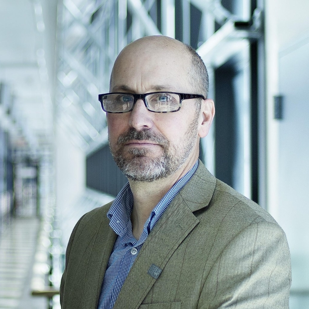 A portrait of a man standing indoors. He has a short beard, wears glasses, and is dressed in a business casual attire with a blue button-up shirt and a khaki blazer. He is standing with his right arm resting on a railing, looking thoughtfully towards the camera with a light-filled, modern corridor with glass panels and metal structures in the background.