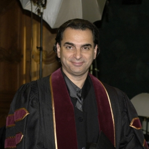 The image shows a man smiling at the camera, wearing a dark academic gown with maroon and gold accents on the sleeves, indicating a ceremonial occasion, possibly a graduation or academic event.