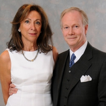 Portrait of a couple: long-haired woman wearing a white sleeveless dress and necklace, and man wearing a dark, 3-piece suit with white shirt and blue tie with white pocket scarf