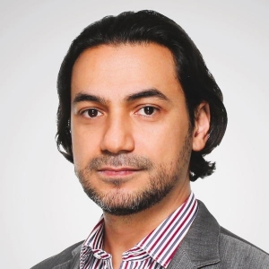 Portrait of a man with nape-length black hair and facial stubble. He is wearing a grey blazer and striped dress shirt.