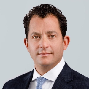 Portrait of a serious-looking, clean-shaven man with short black hair. He is wearing a business suit with a white shirt and blue tie.