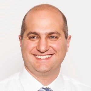 A man is smiling and wearing a white collared shirt with grey striped tie.