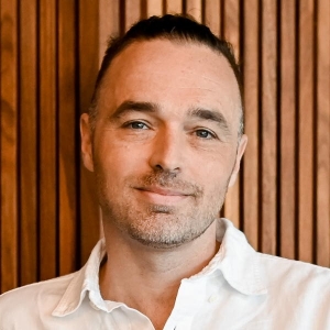 A man with short dark hair is wearing a white collared shirt and is standing in front of a wooden panel wall.